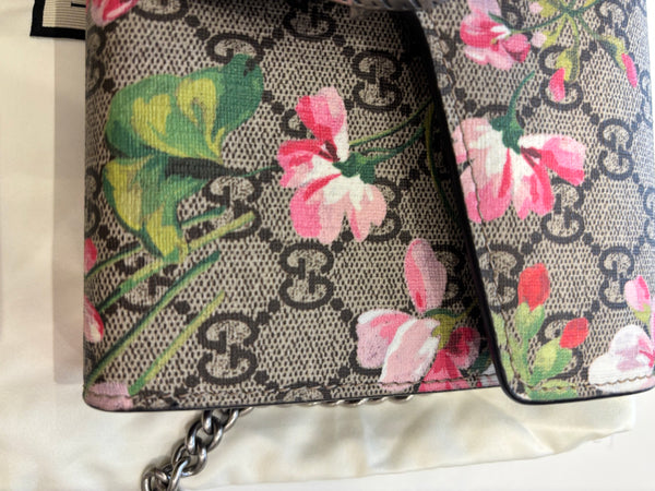 A close-up of a Gucci shoulder bag featuring the brand's classic GG Supreme canvas in beige and brown, decorated with vibrant pink and green floral and leaf patterns. A silver chain strap is visible at the bottom left.
